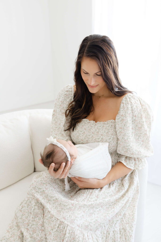 A smiling new mother gazes down to her newborn in her hands while sitting on a white couch after visiting the Jacksonville tongue tie center