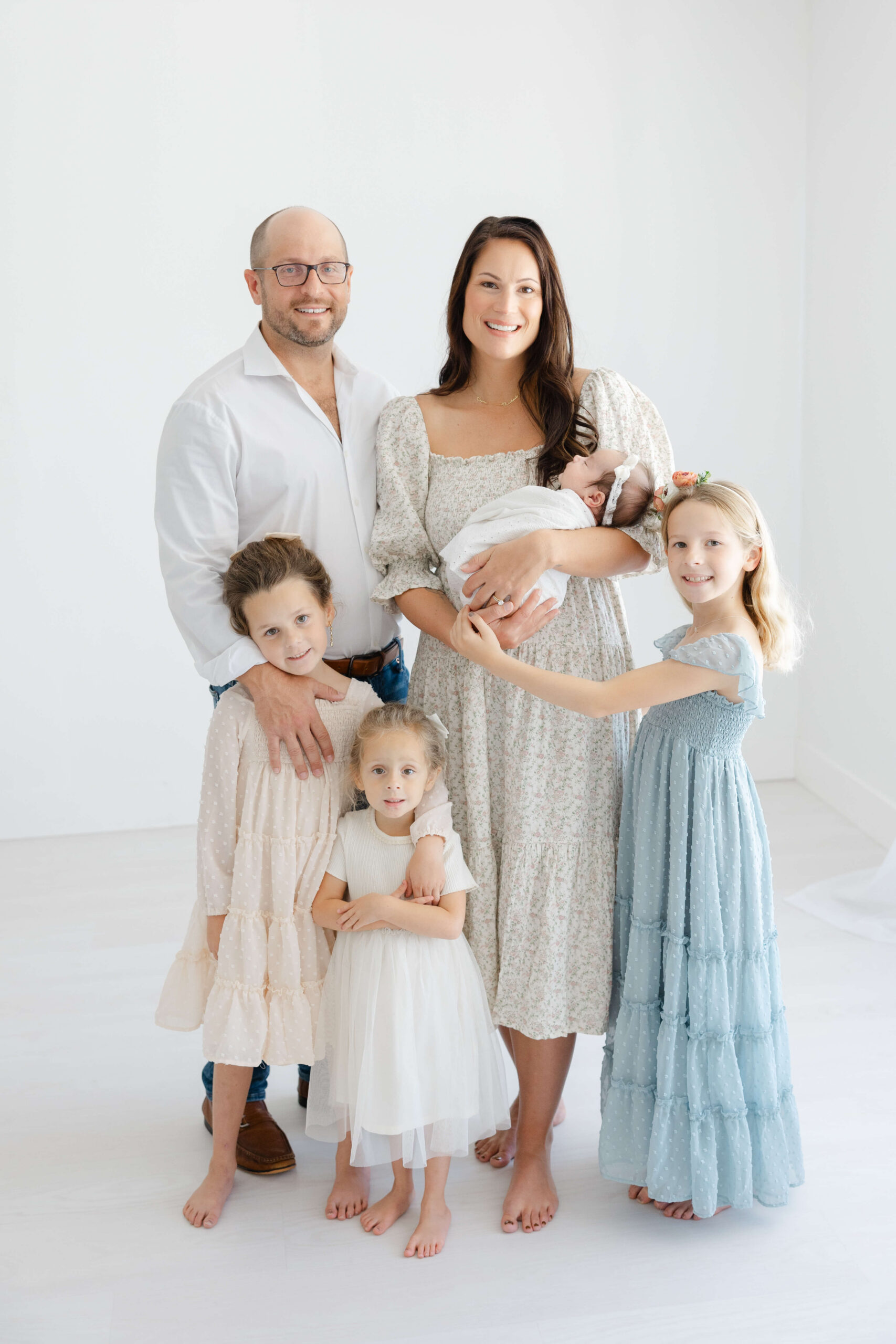 A mom and dad stand holding their newborn as their three toddler daughters in dresses smile around them in a studio after visiting the Jacksonville tongue tie center