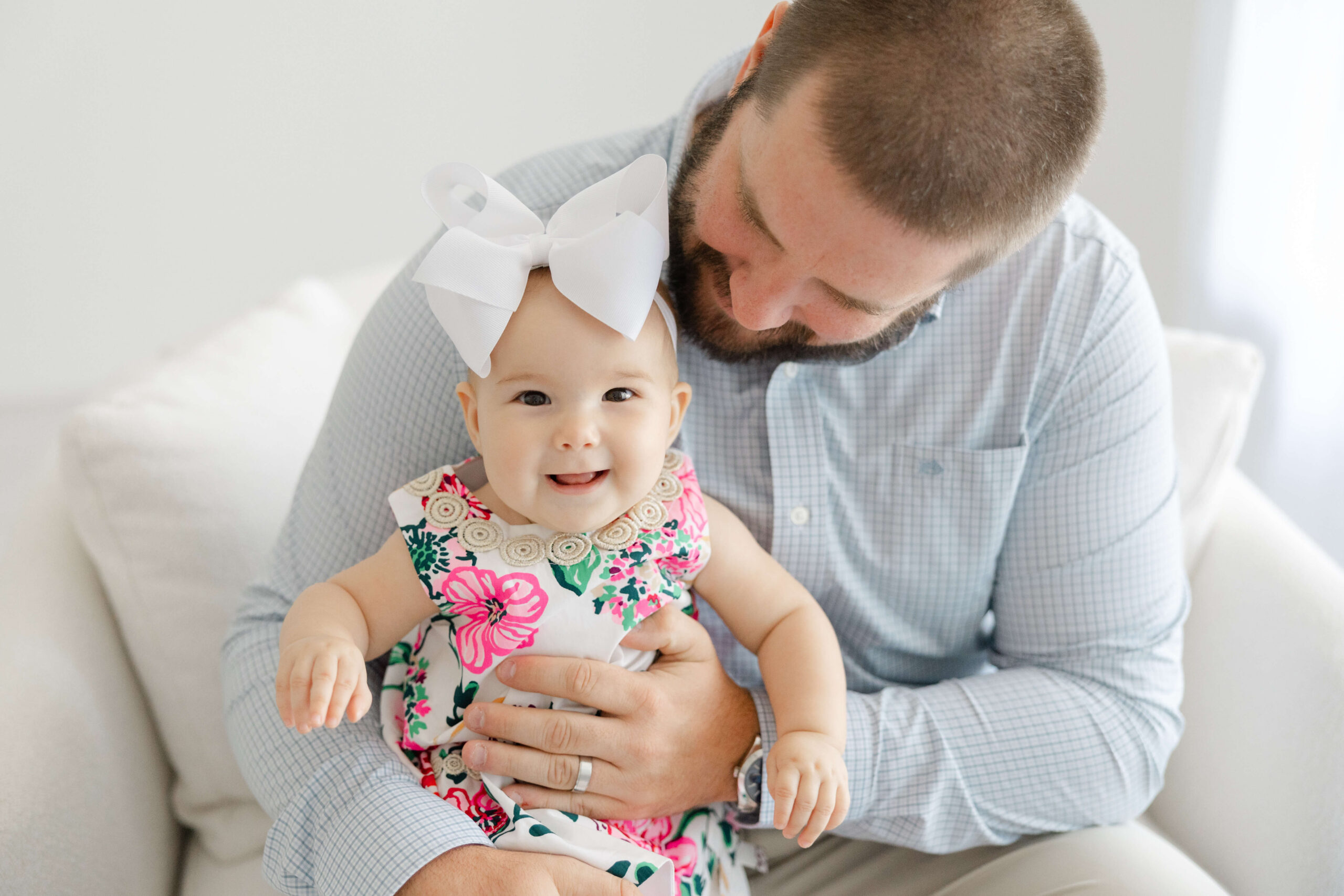 A smiling baby girl sits in dad's lap in a pink flower dress in a chair under a window