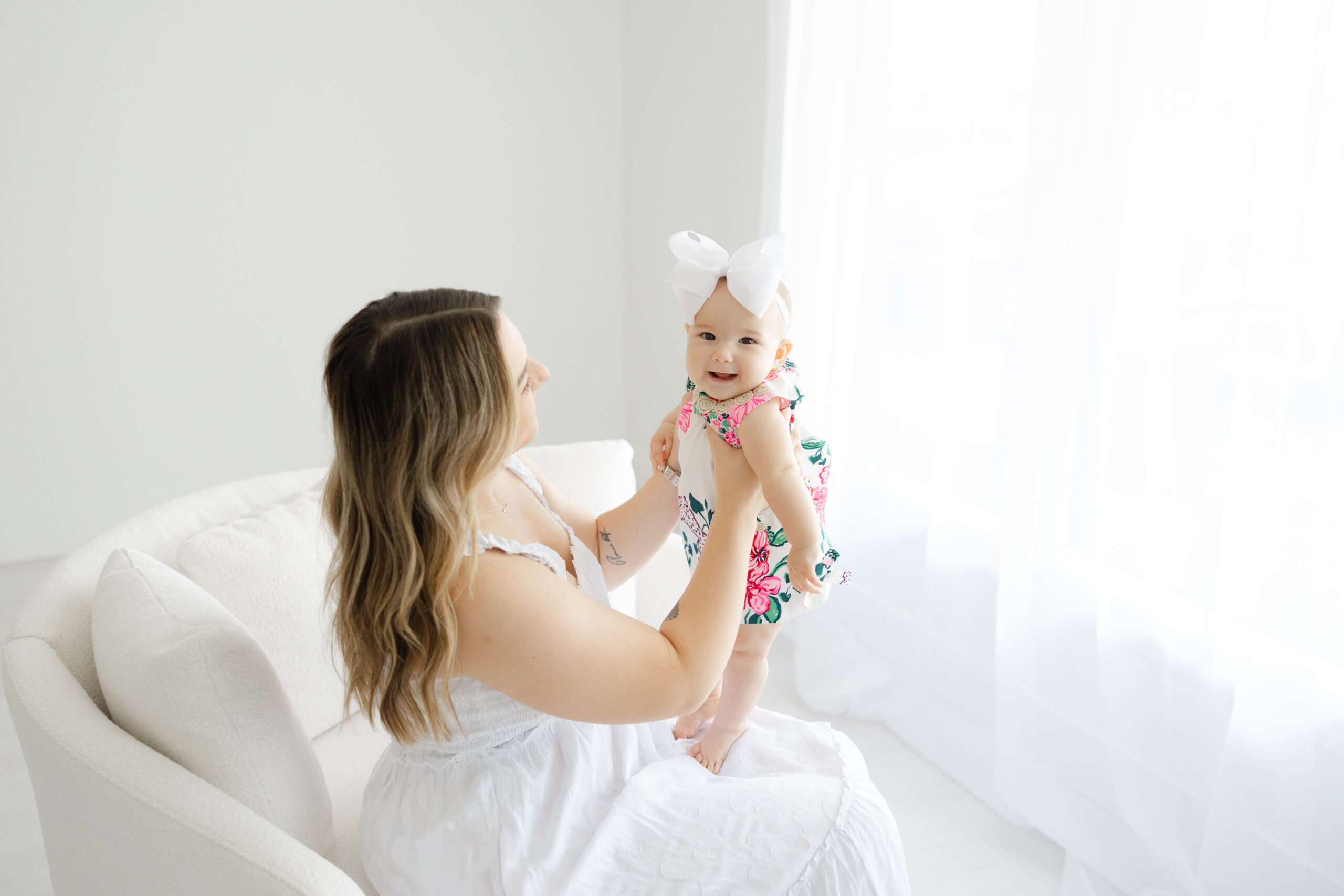 A happy baby girl in a pink flower dress smiles while playing with mom in a chair after meeting an au pair in jacksonville fl