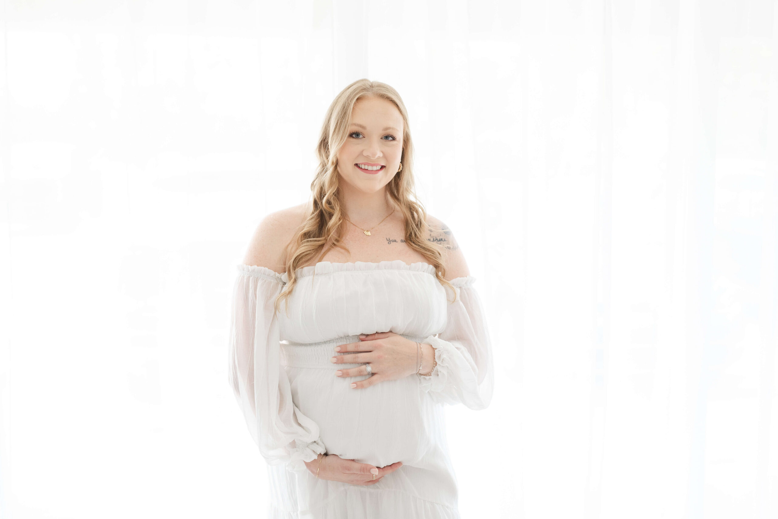 A smiling mother to be standing in a studio in a white maternity gown with hands on her bump