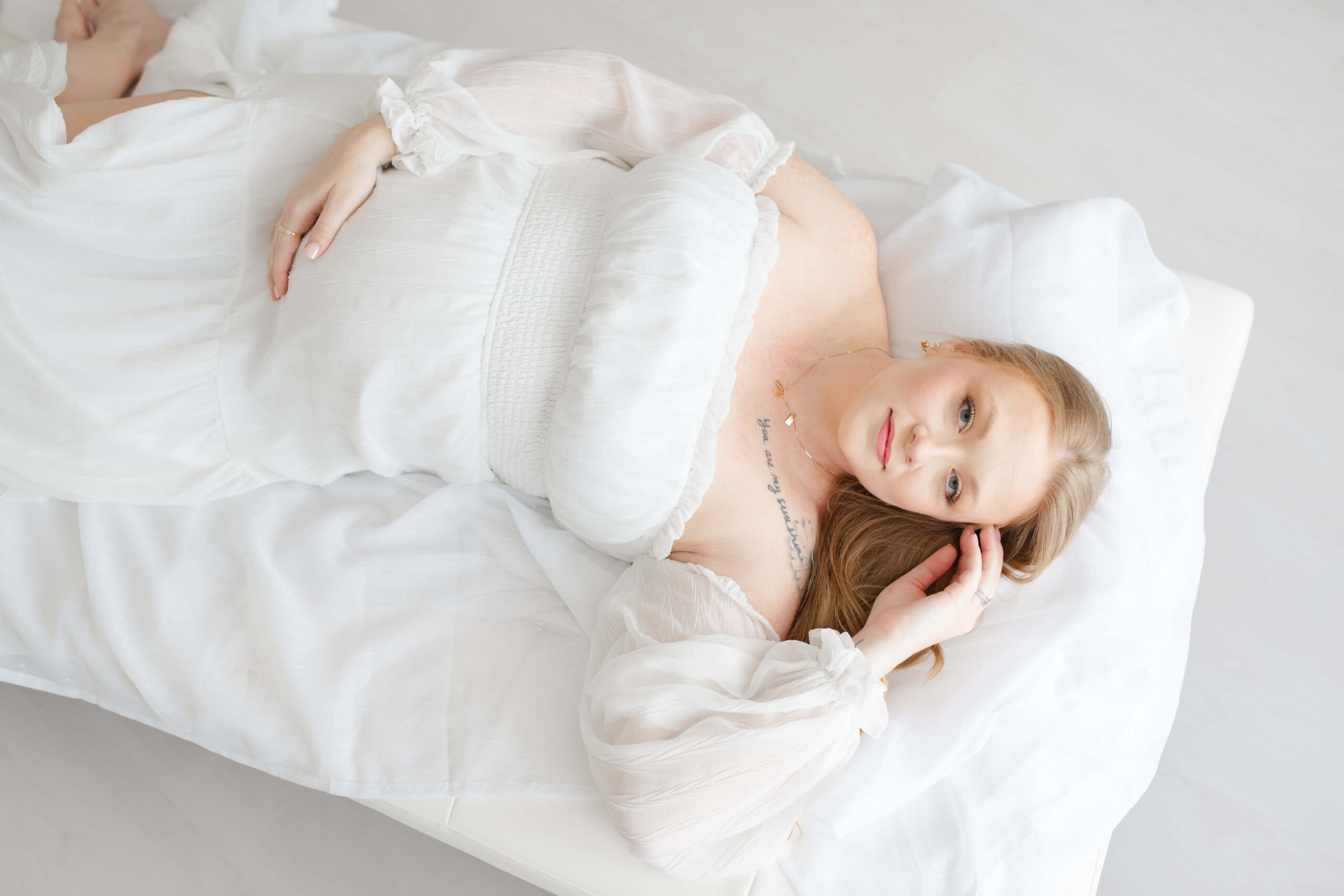 A mother to be lays across a couch in a white maternity gown with a smile