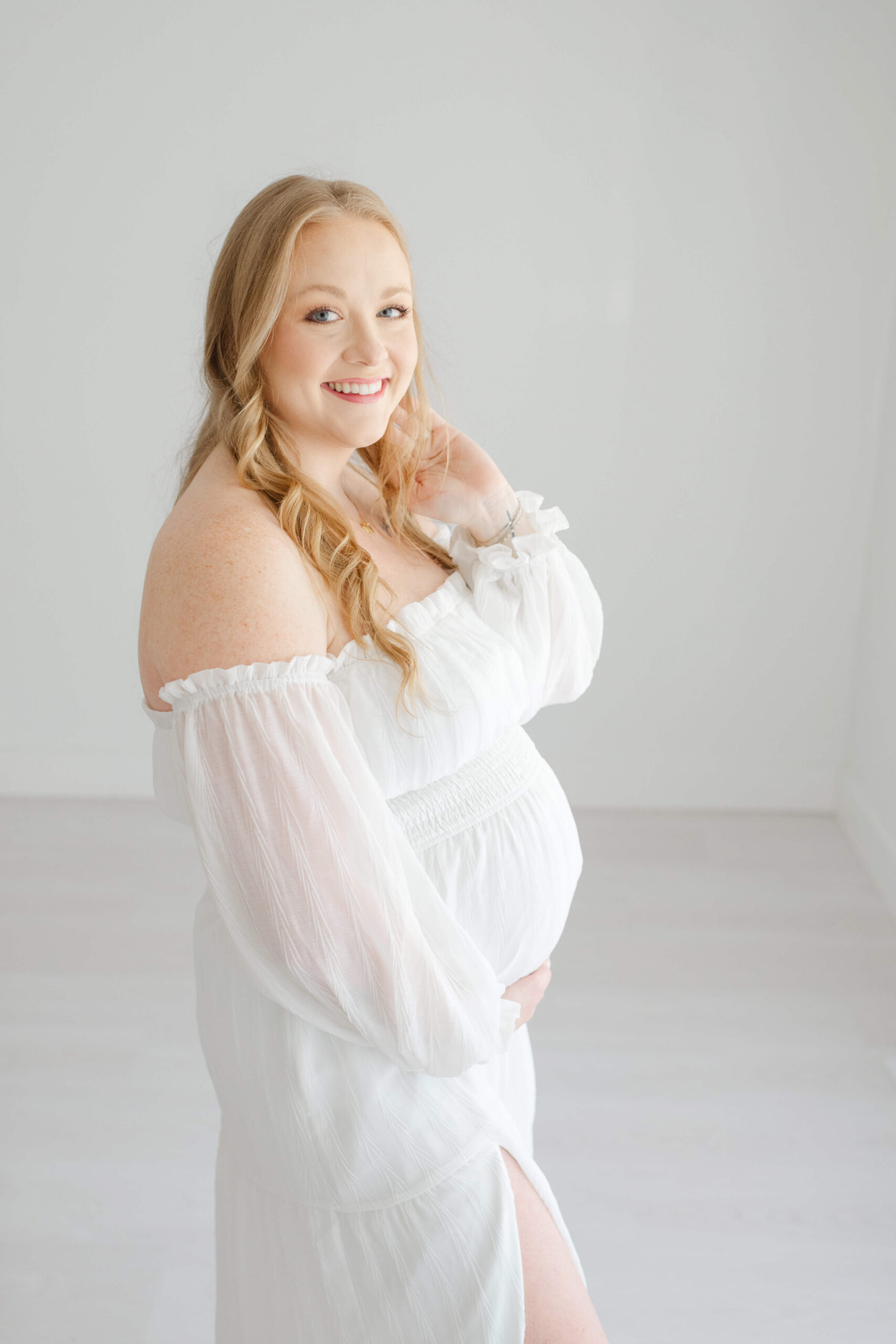 A happy expecting woman stands in a studio with a hand in her hair and under her bump in a white maternity dress after meeting birthing with jane