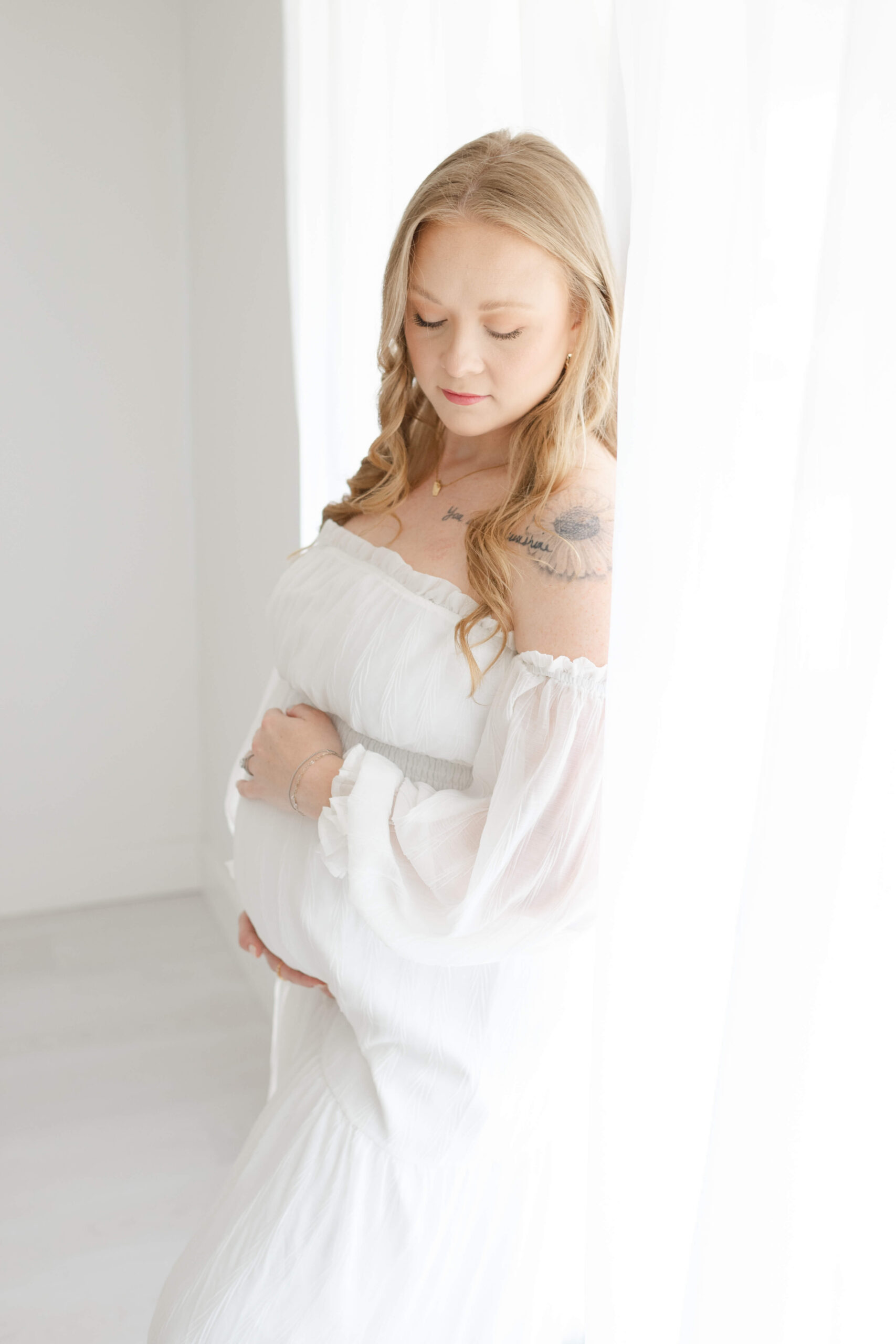 A pregnant woman stands in a window gazing down to her bump with hands on top and bottom after meeting birthing with jane