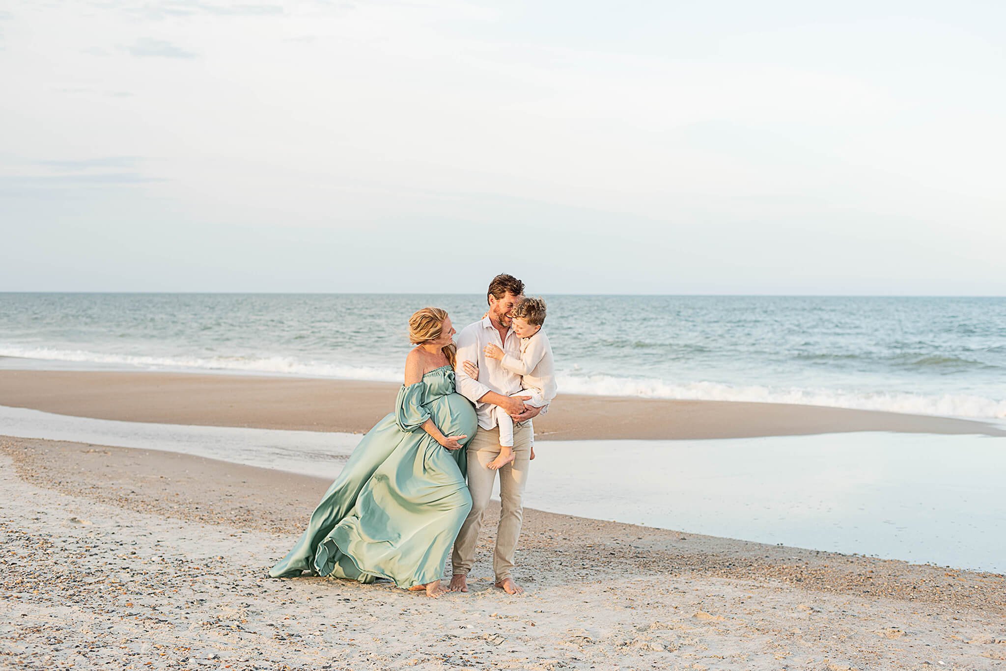 A happy mom and dad laugh with their toddler son on dad's hip as they stand on a beach at sunset after visiting blooming bellies jacksonville
