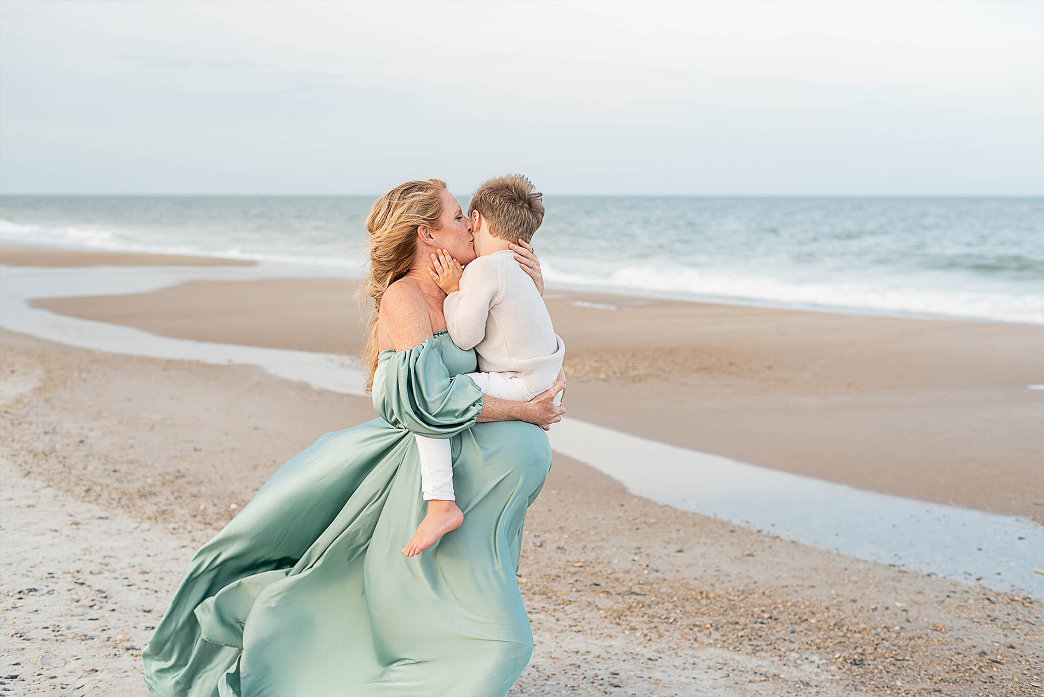 A pregnant mom kisses her toddler son on a beach as he sits on her bump