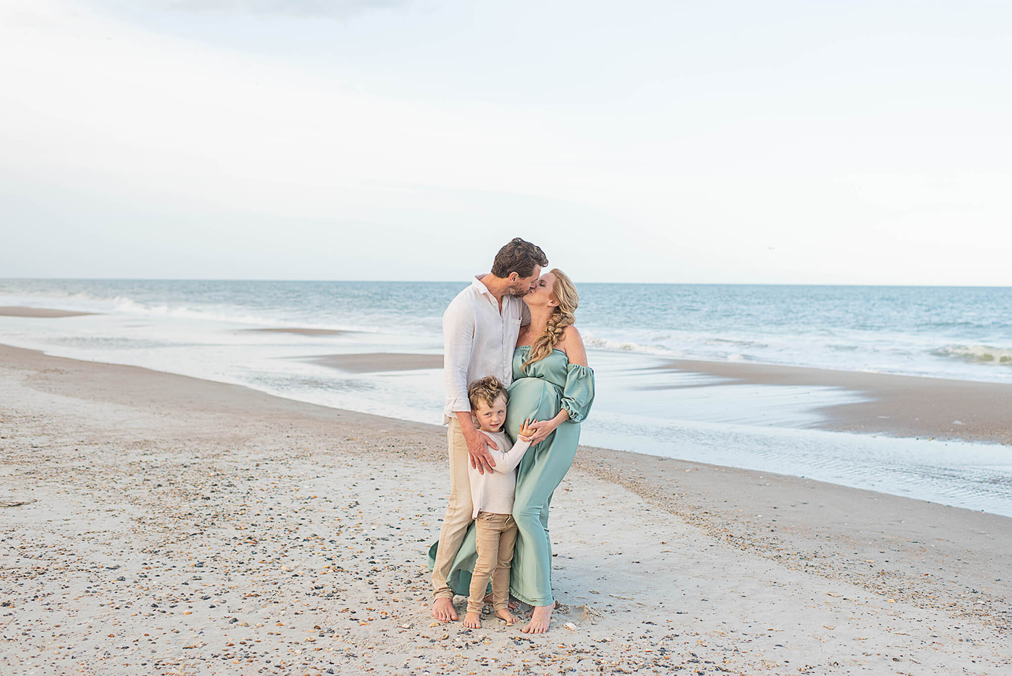 A happy expecting couple kisses on a beach as their toddler son hugs mom's bump