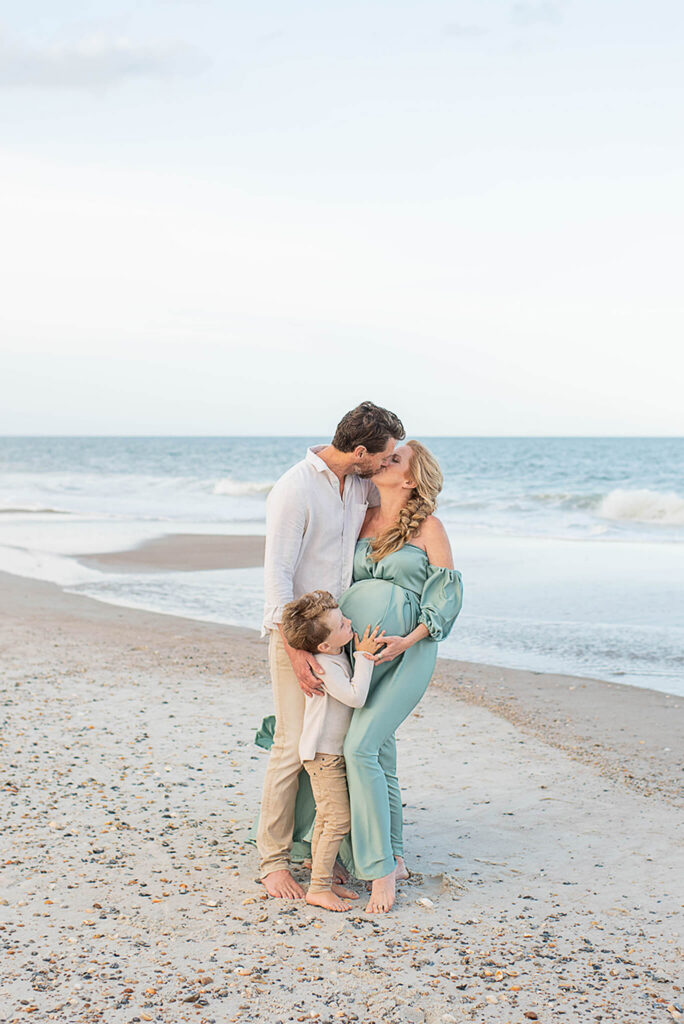 Mom and dad kiss as toddler son hugs mom's pregnant belly on a beach at sunset in white and green after visiting blooming bellies jacksonville