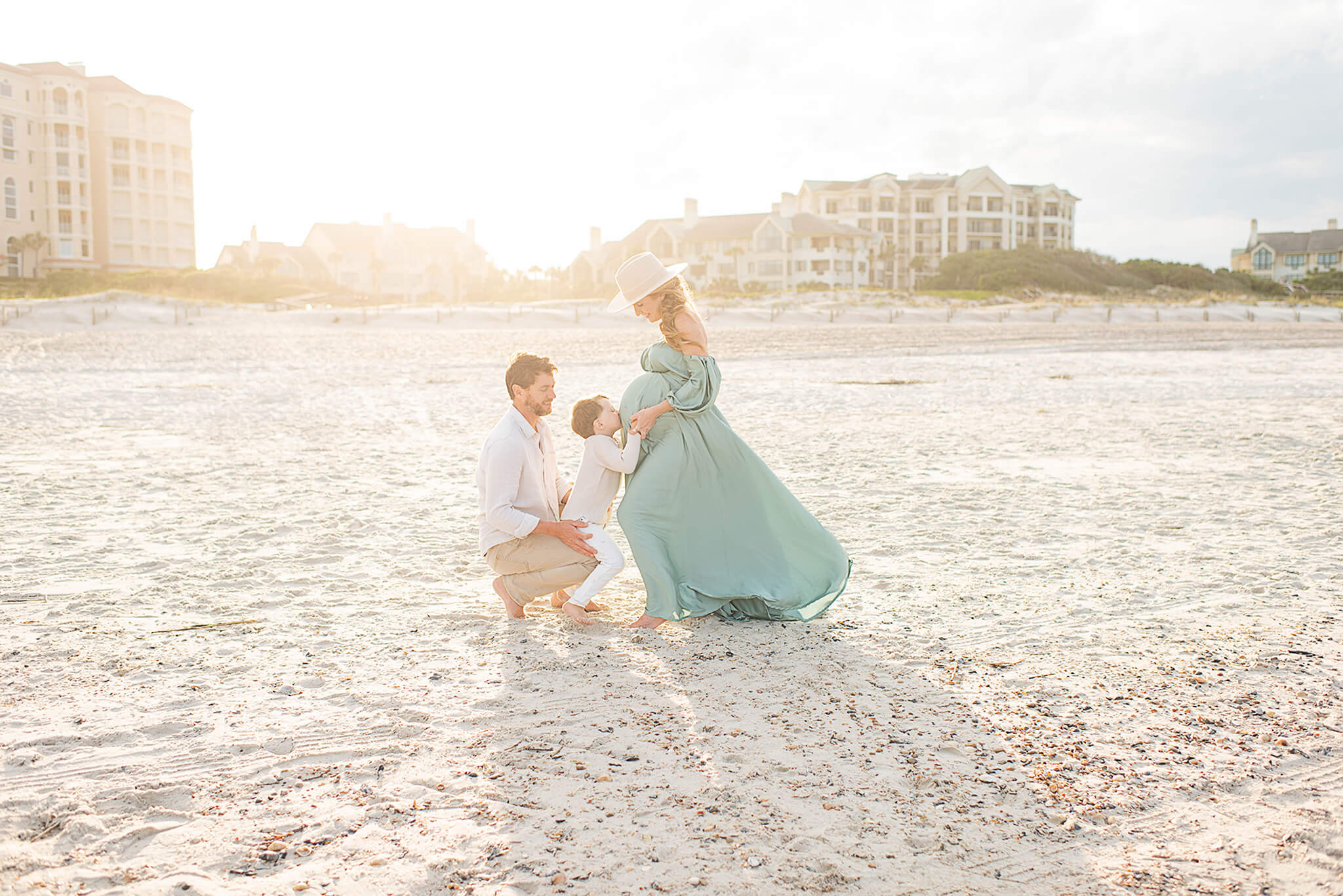 A toddler boy kisses mom's bump while on a beach at sunset ina. green maternity gown and large hat after visiting blooming bellies jacksonville
