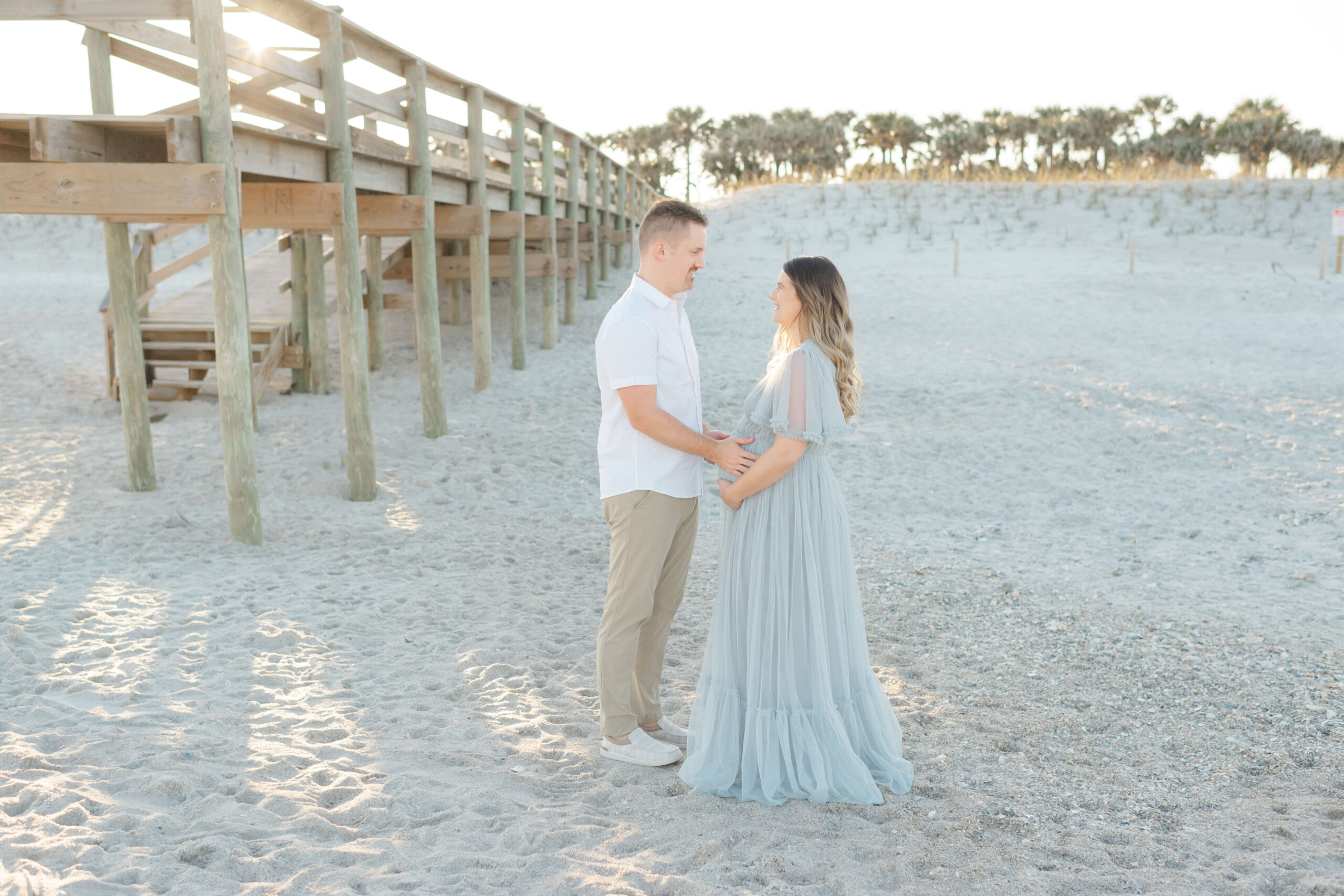 A happy expecting couple stands on a beach at sunset smiling at each other