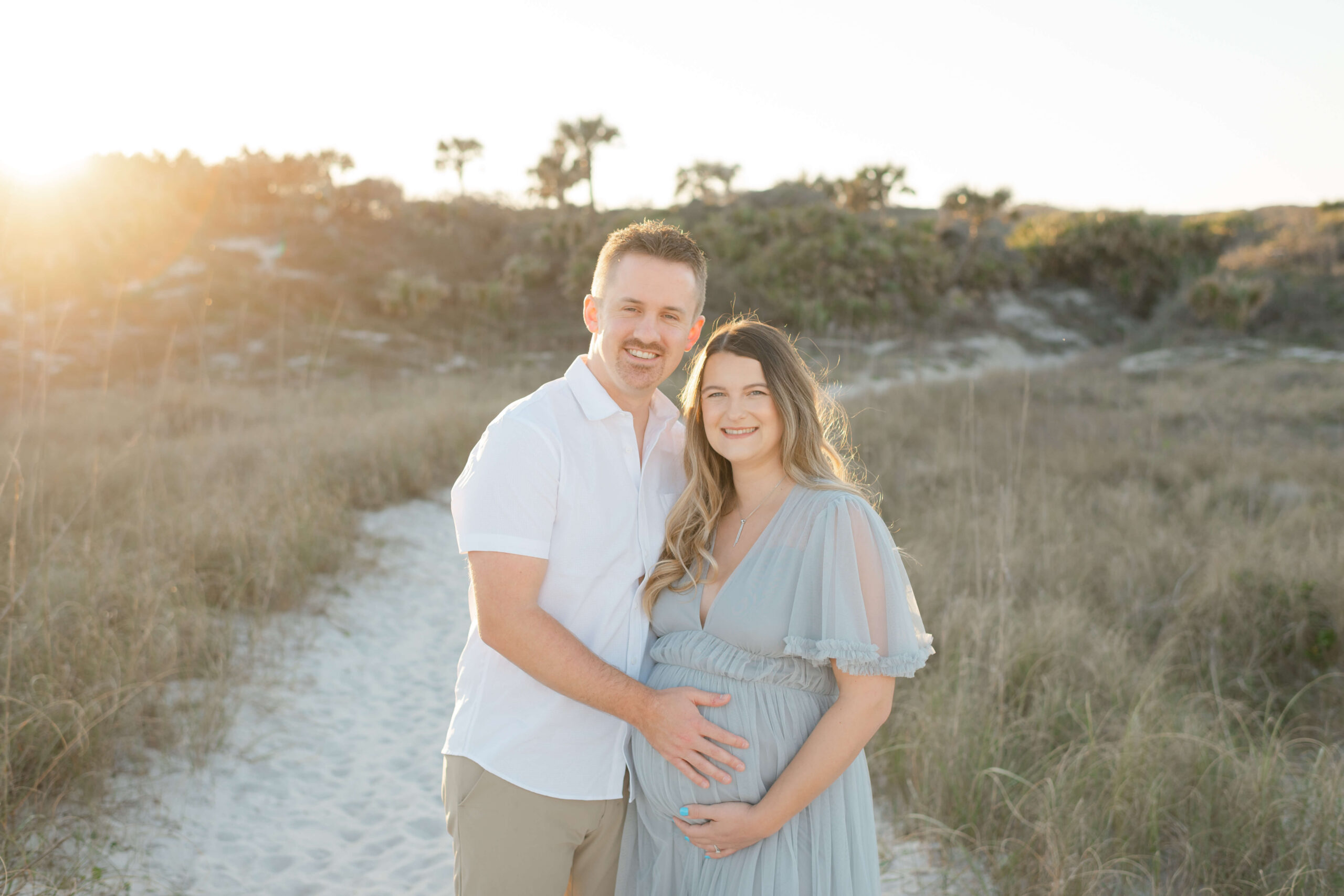 A happy pregnant couple stands on a beach trail through the dune at sunset smiling in blue and white before heading to date night ideas in jacksonville fl