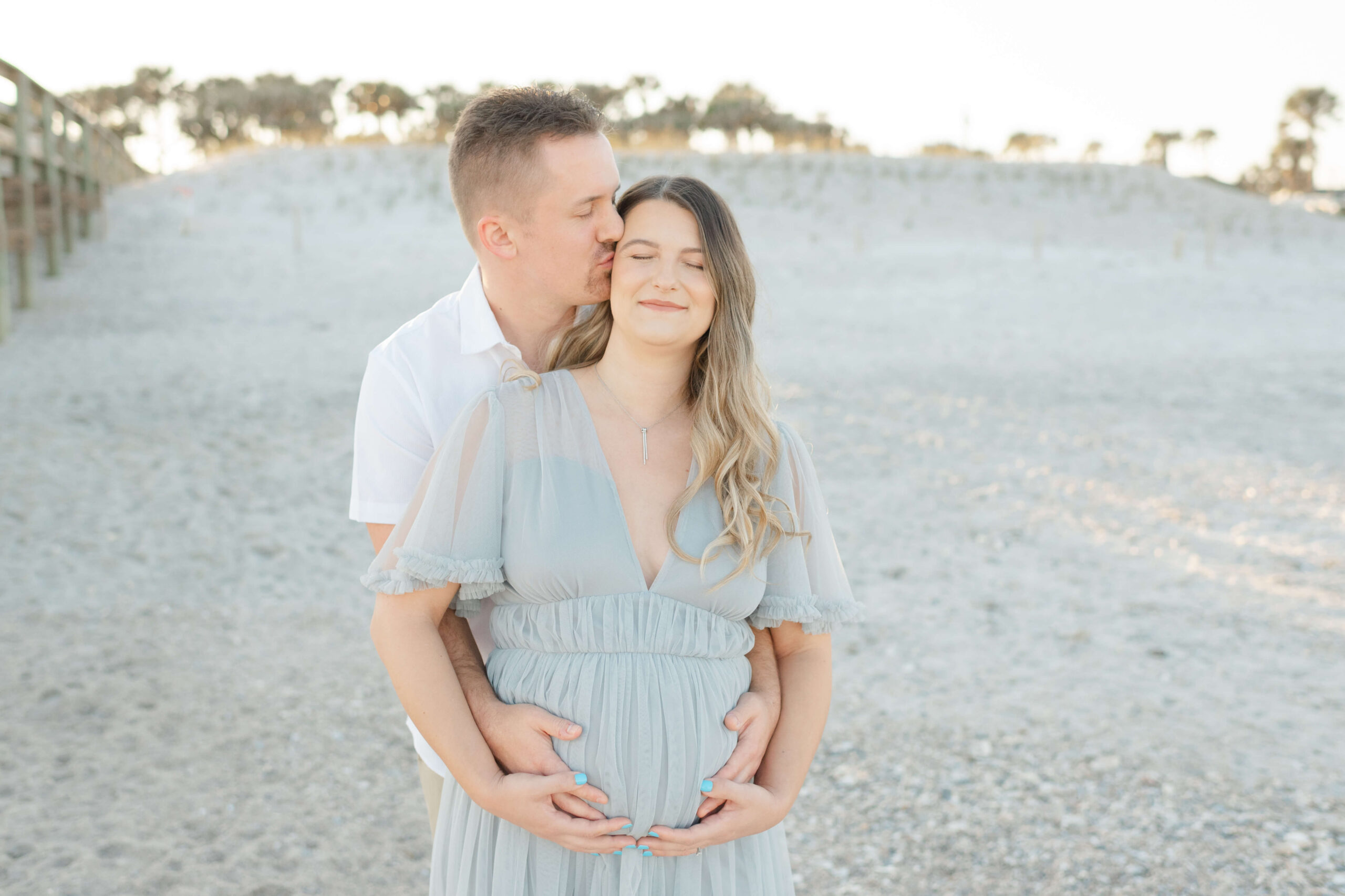 A pregnant couple snuggles and kisses on a beach at sunset with hands under the bump before heading to date night ideas in jacksonville fl