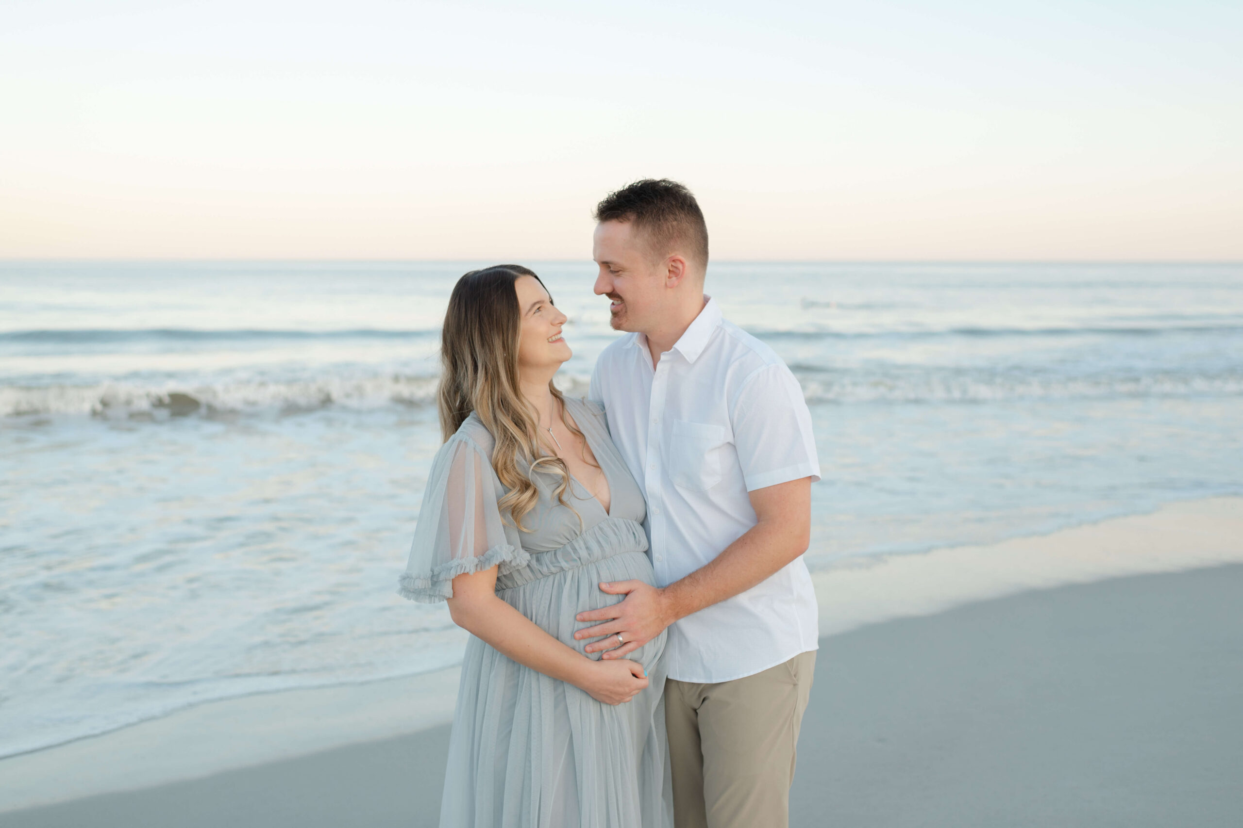 A happy expecting couple stands on a beach at sunset with hands on the bump in a blue maternity gown after finding date night ideas in jacksonville fl
