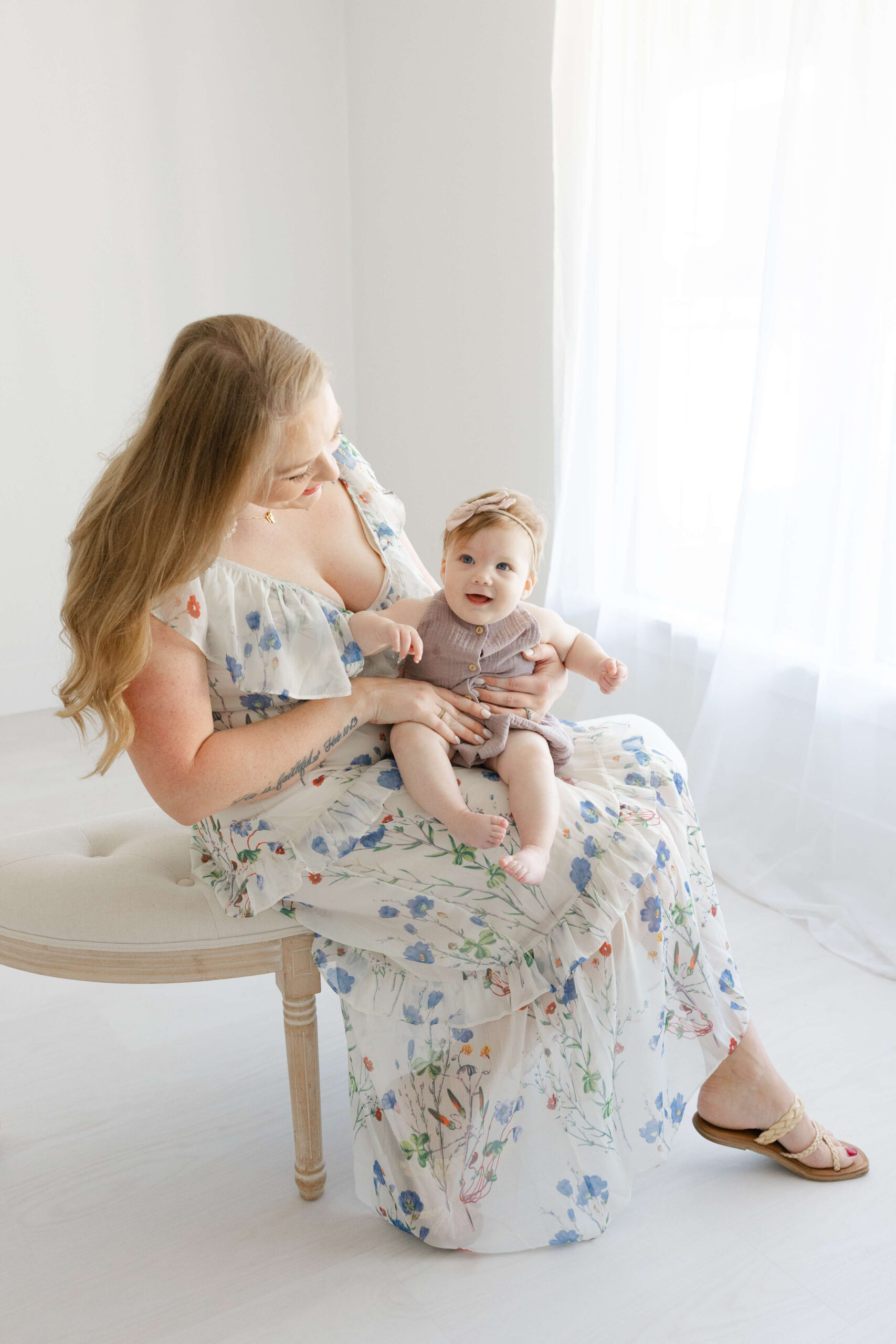 A happy mom in a floral print dress plays with her baby daughter in her alp on a bench in a studio