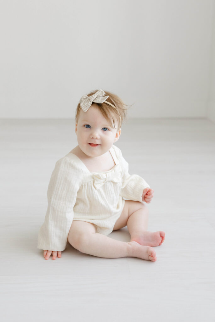 Infant photography outfit ideas with a sitting baby wearing a simple cream romper and neutral bow in a bright studio.