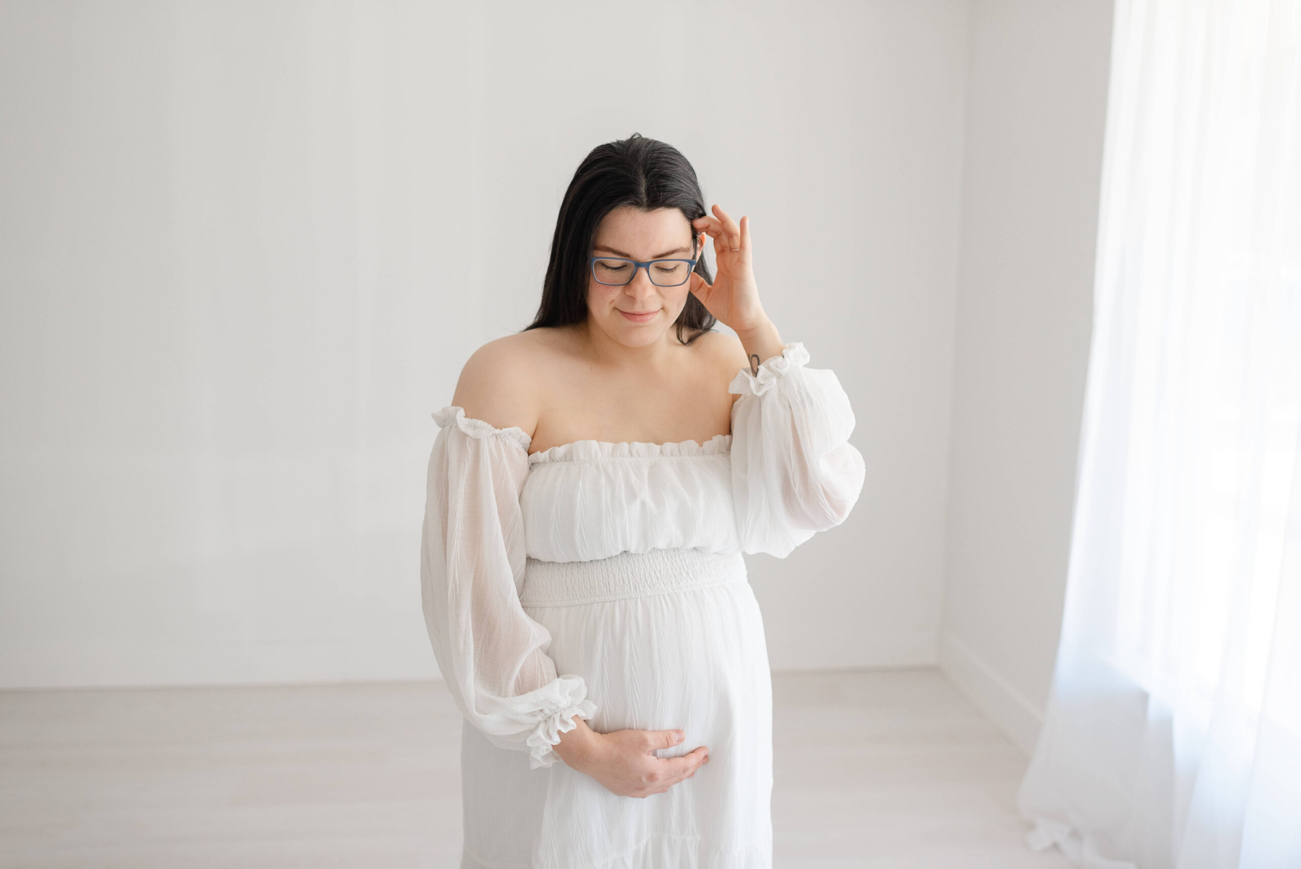 A mom to be in a white maternity gown stands in a studio holding her bump