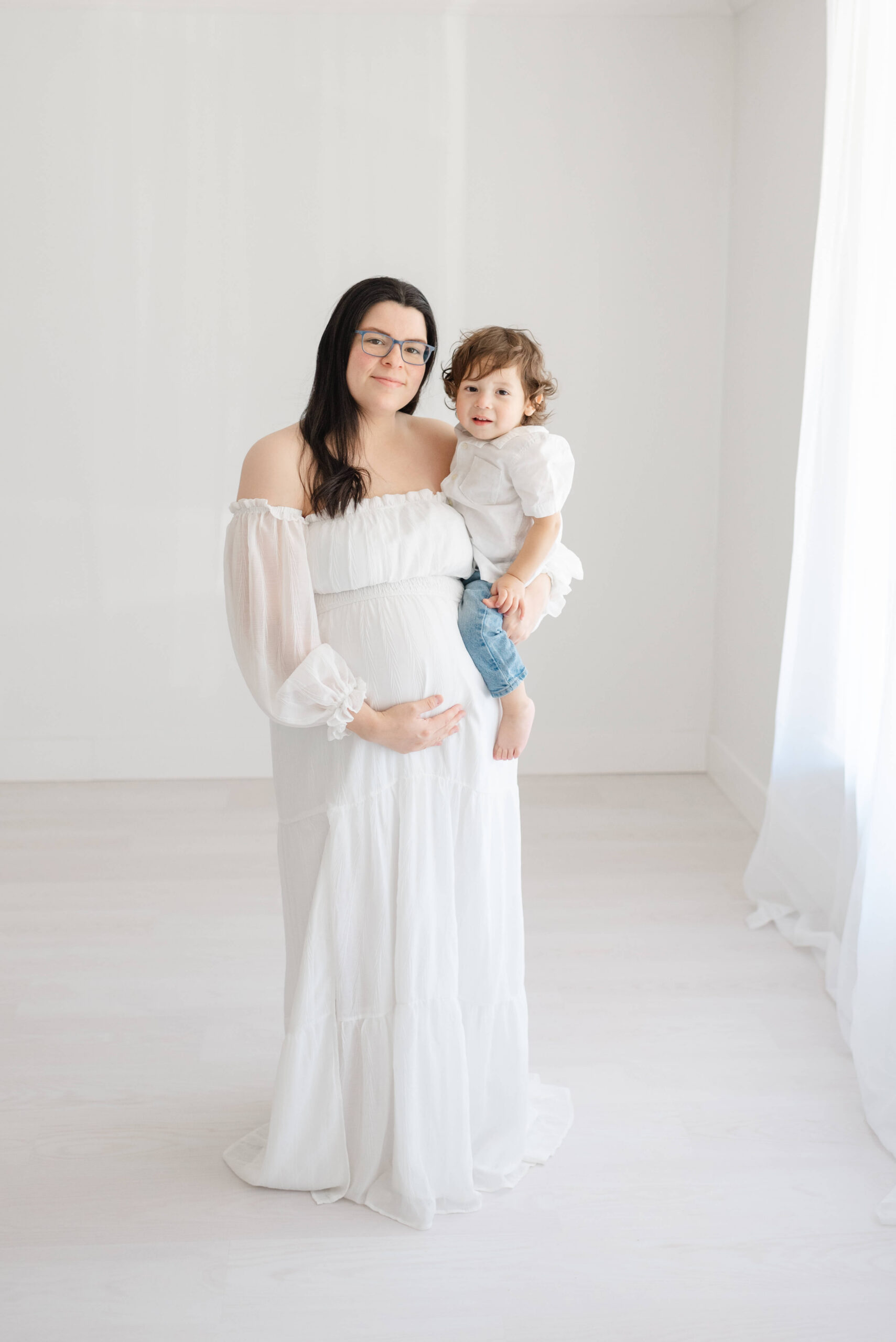 A smiling pregnant mother stands in a window holding her toddler son on her hip