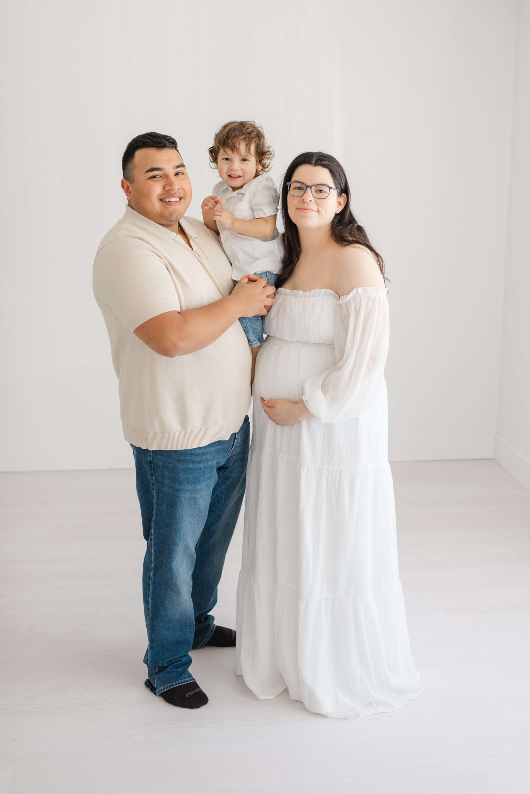 A happy expecting mom and dad stand in a studio holding their toddler son in white and denim after enjoying their maternal fetal medicine in jacksonville fl