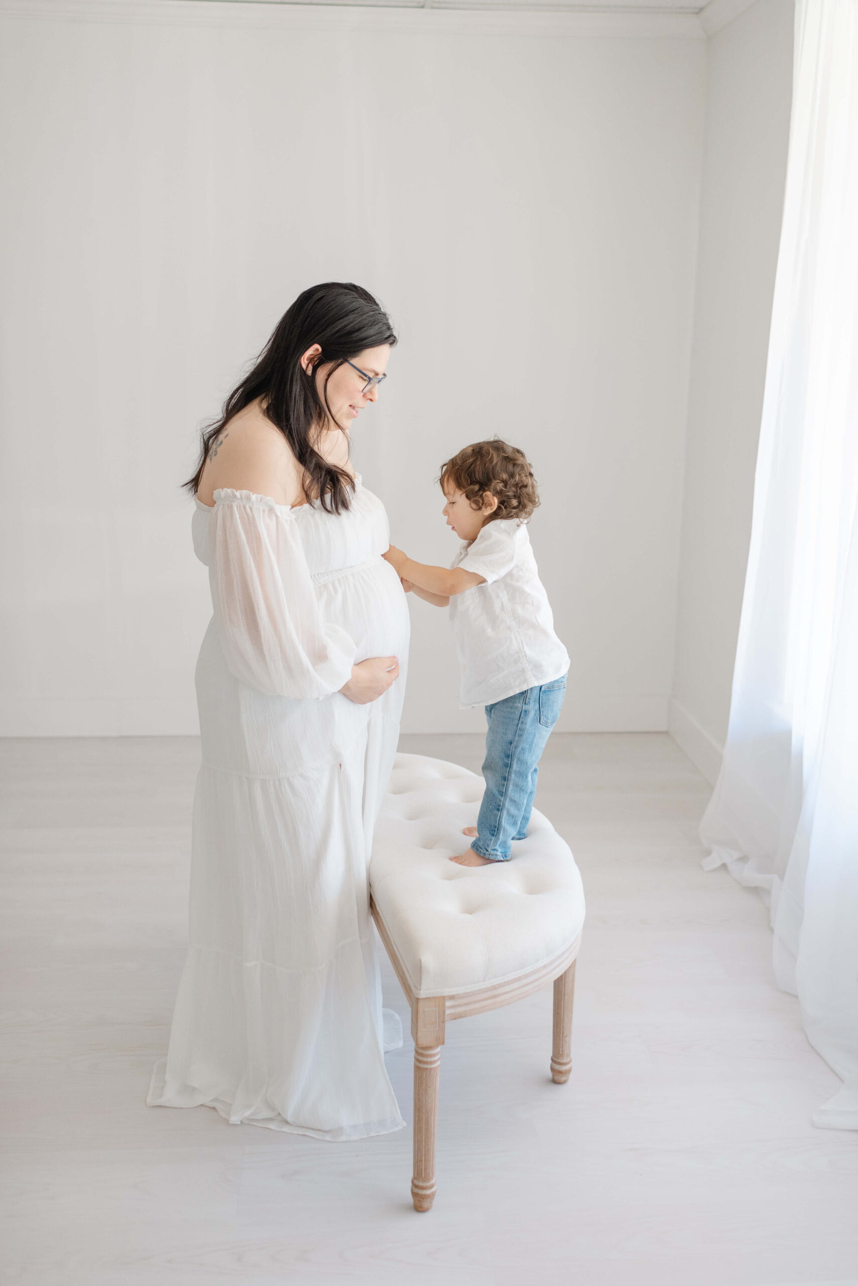 A toddler stands on a bench in a studio while exploring mom's pregnant bump in a white shirt after finding maternal fetal medicine in jacksonville fl