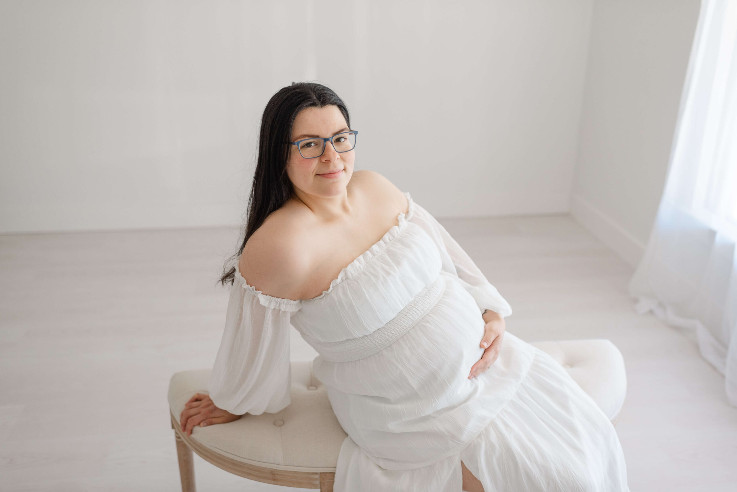 A mother to be in a white maternity gown sits across a bench in a studio under a window after finding maternal fetal medicine in jacksonville fl