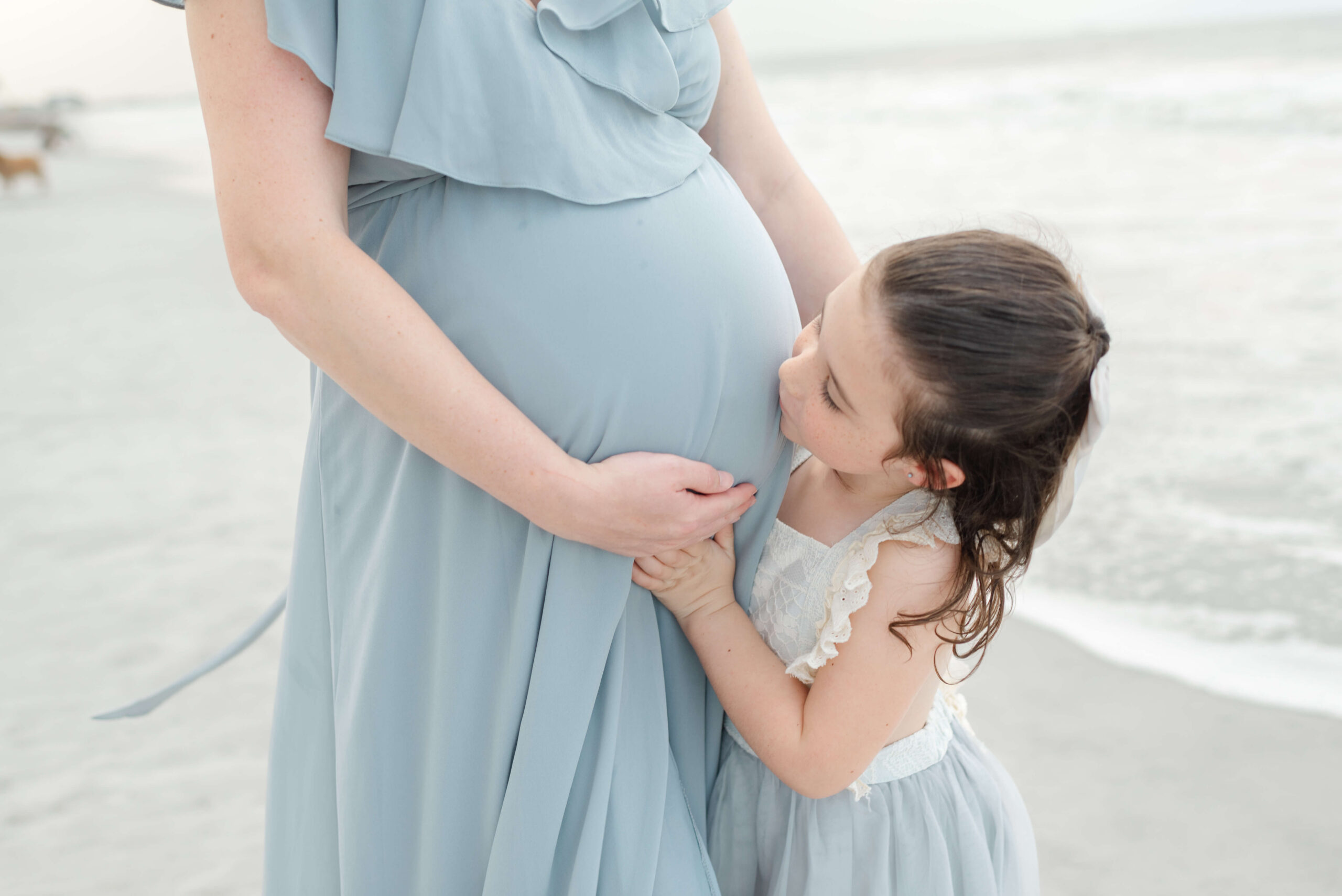 A young girl kisses the bump of her pregnant mother both in blue dresses on a beach