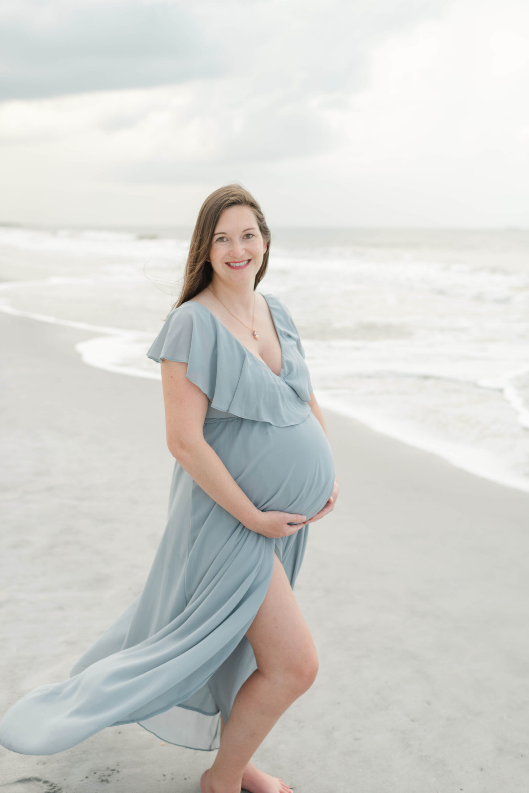 A smiling mother to be walks on a windy beach at sunrise holding her bump in a blue maternity dress