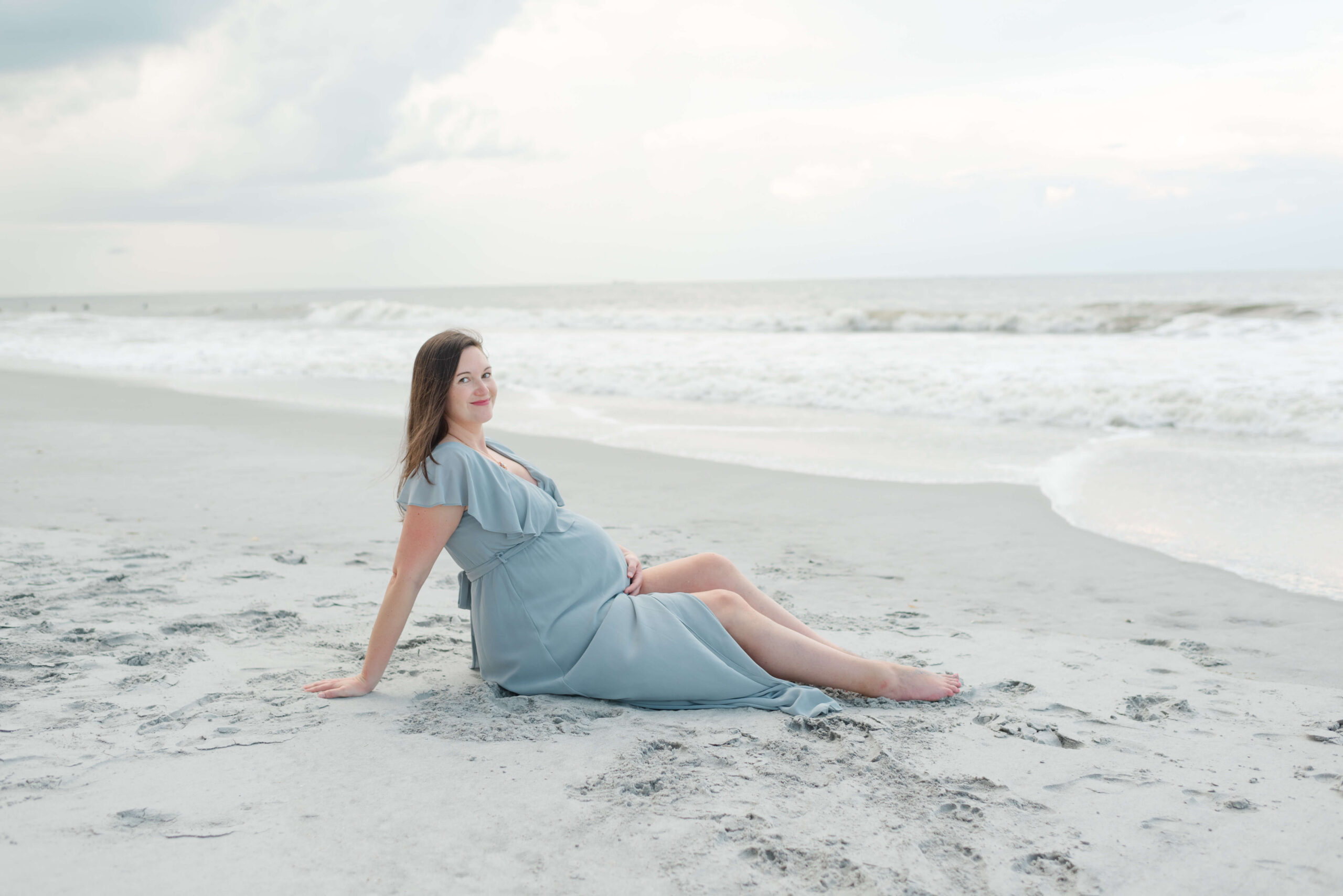 A pregnant woman sits on the beach smiling holding her bump in a blue maternity gown at sunrise after meeting postpartum doulas in jacksonville fl