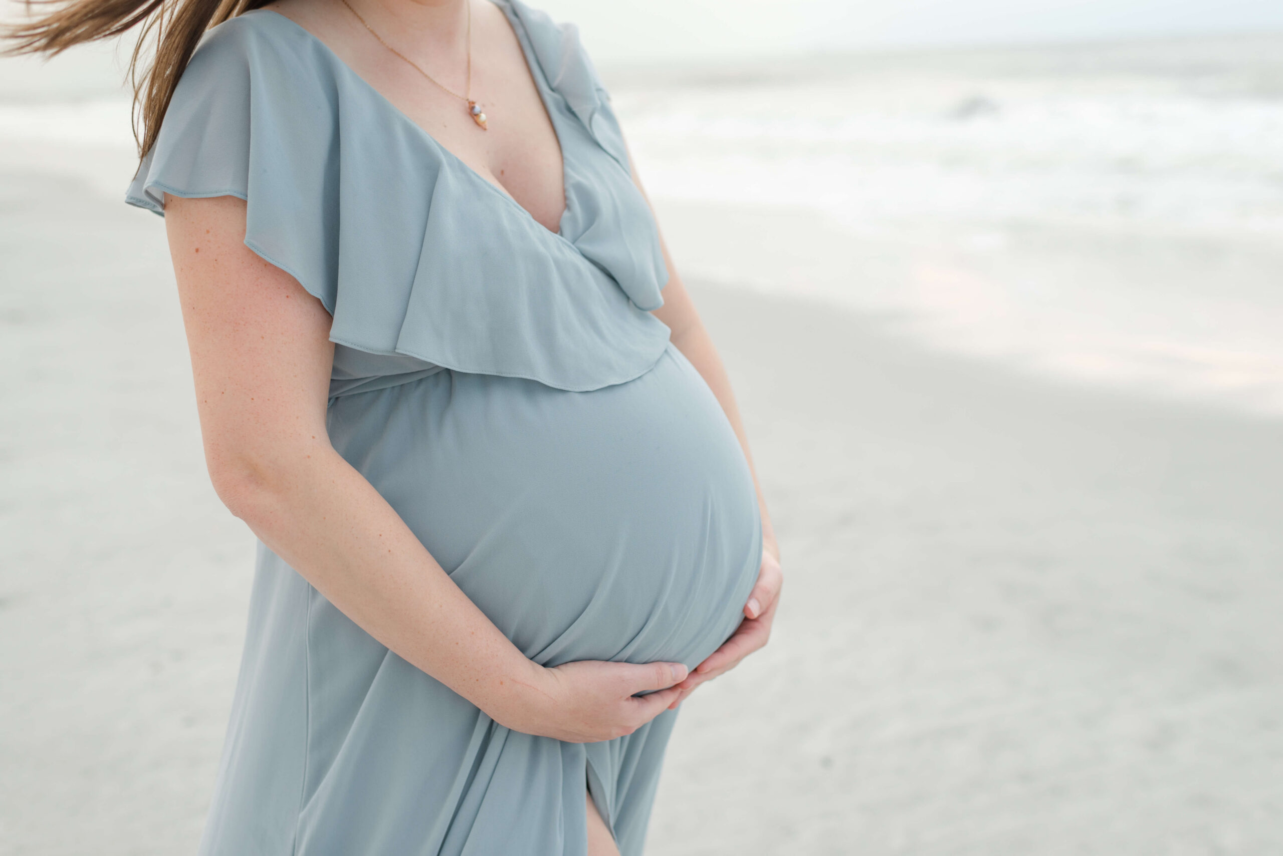 Details of a mother to be's bump while standing on a beach in a blue maternity gown