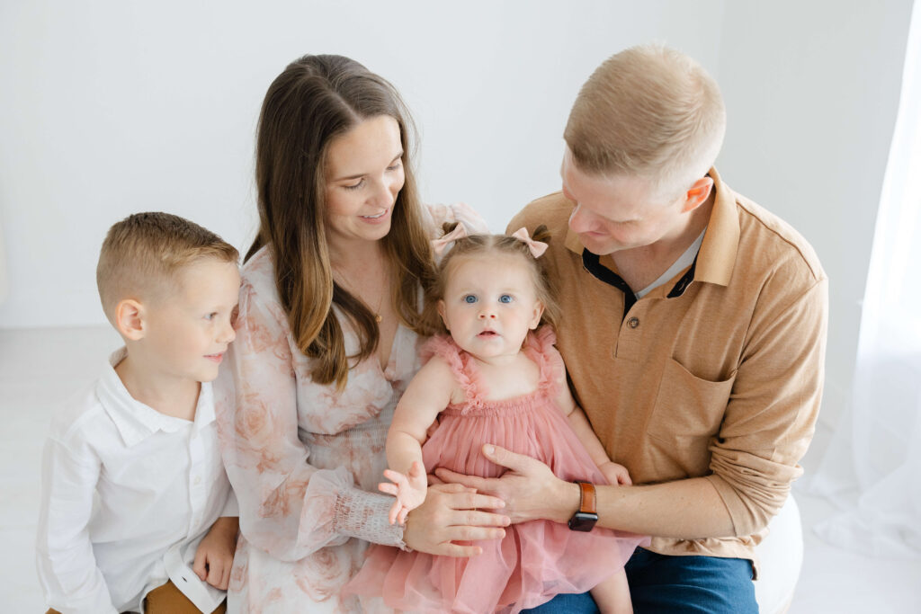 Happy mom and dad sit on a bench with their toddler in a pink dress as bigger brother looks on