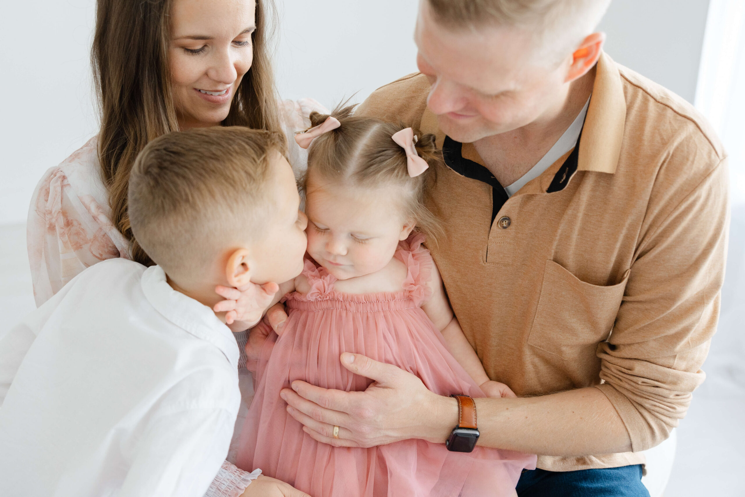 A toddler boy leans in to kiss younger sister sitting in dad's lap in a pink dress after visiting preschools in jacksonville fl