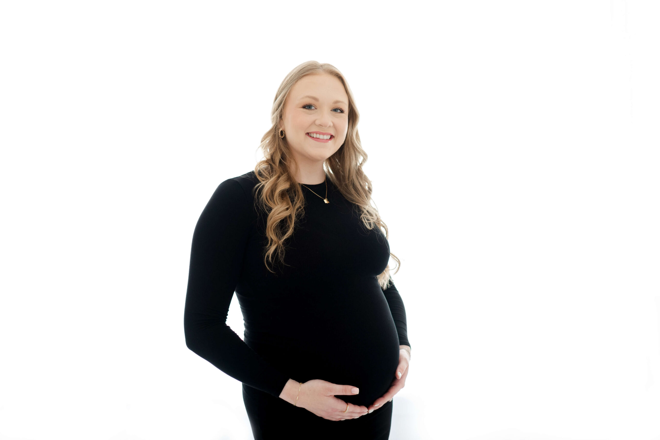 A smiling pregnant woman with her hands under her bump while standing in a studio