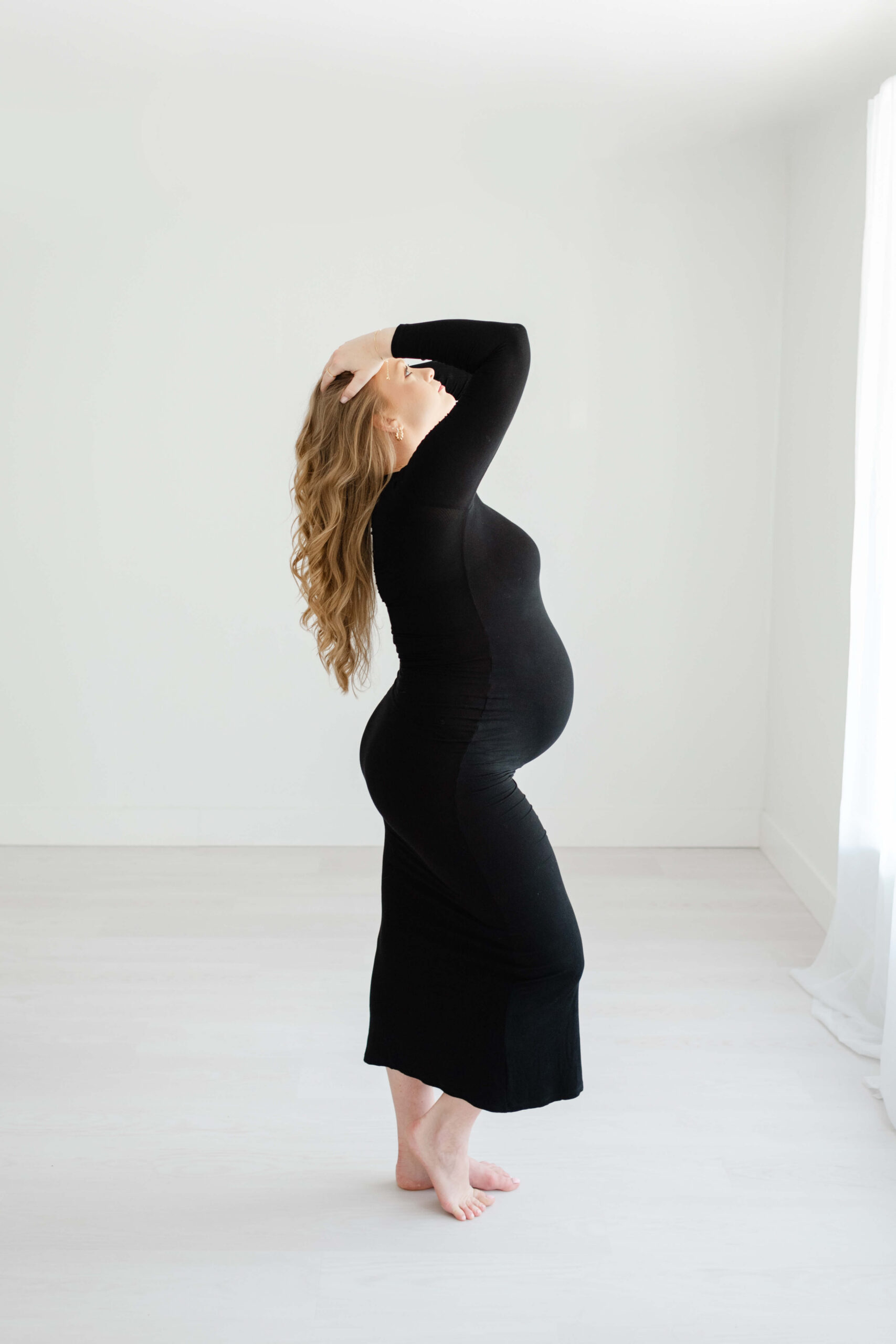 A pregnant woman in a black gown stands in a studio with hands in her hair after visiting srb obgyn