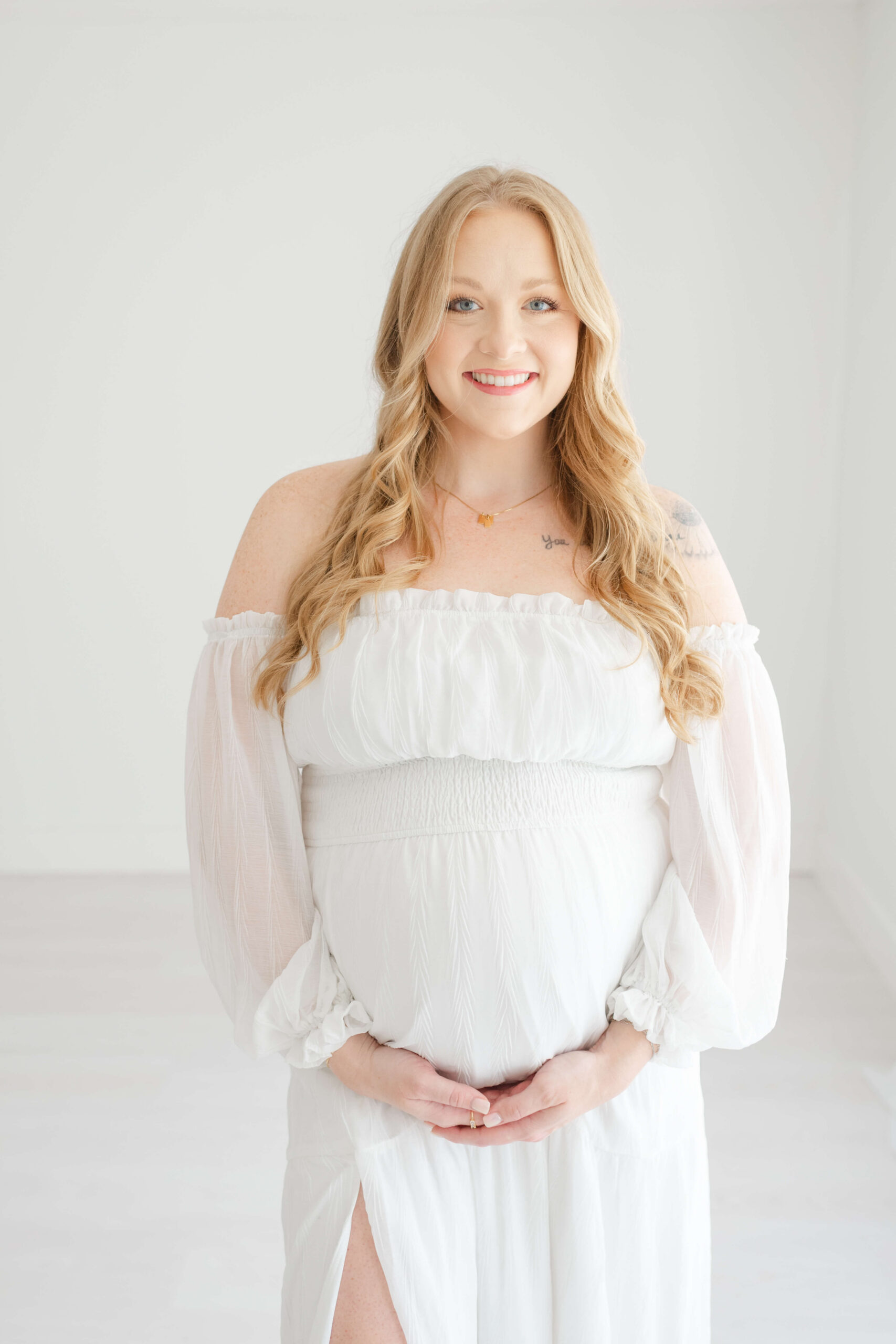 A happy mother to be stands smiling in a white maternity dress and hands under her bump after visiting srb obgyn