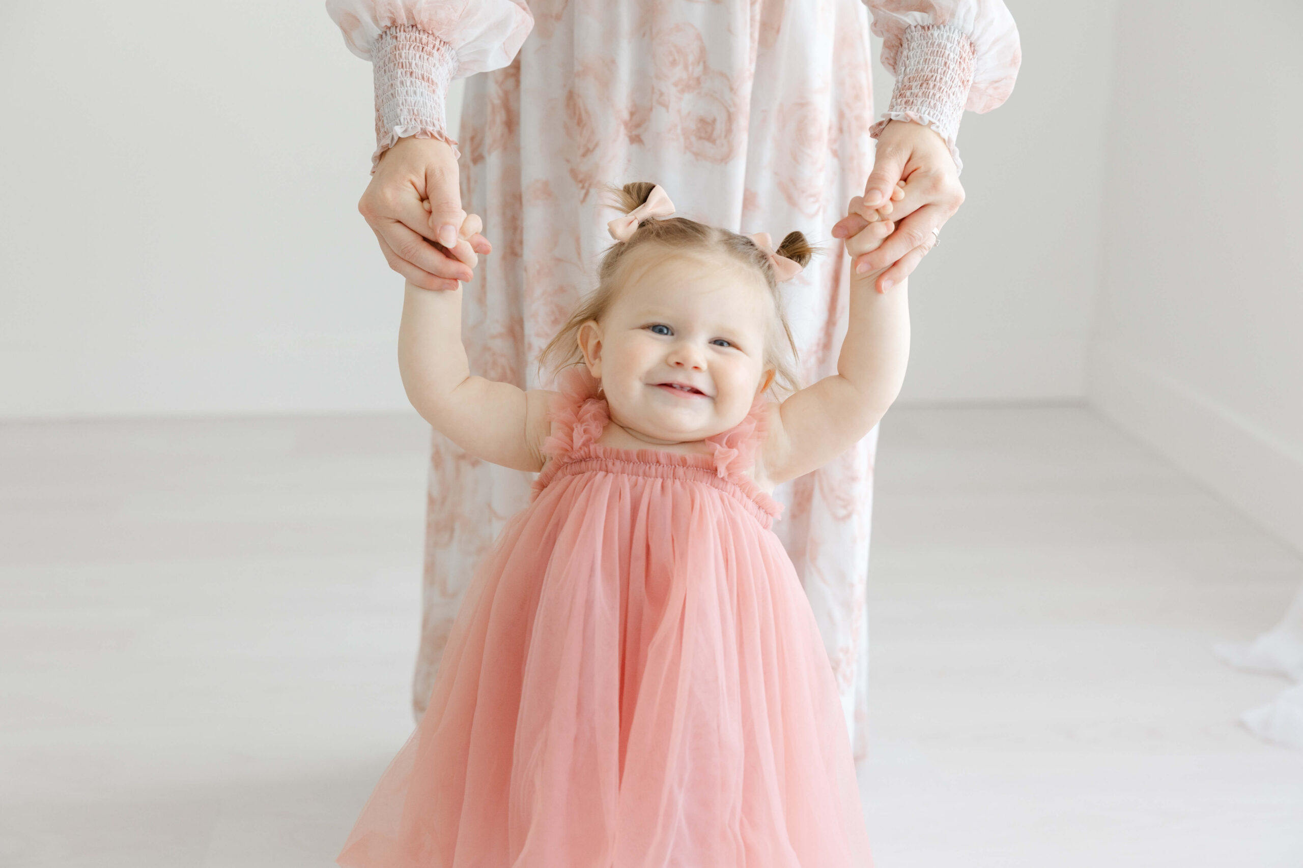 A smiling toddler girl in a pink dress walks holding mom's hands in a studio