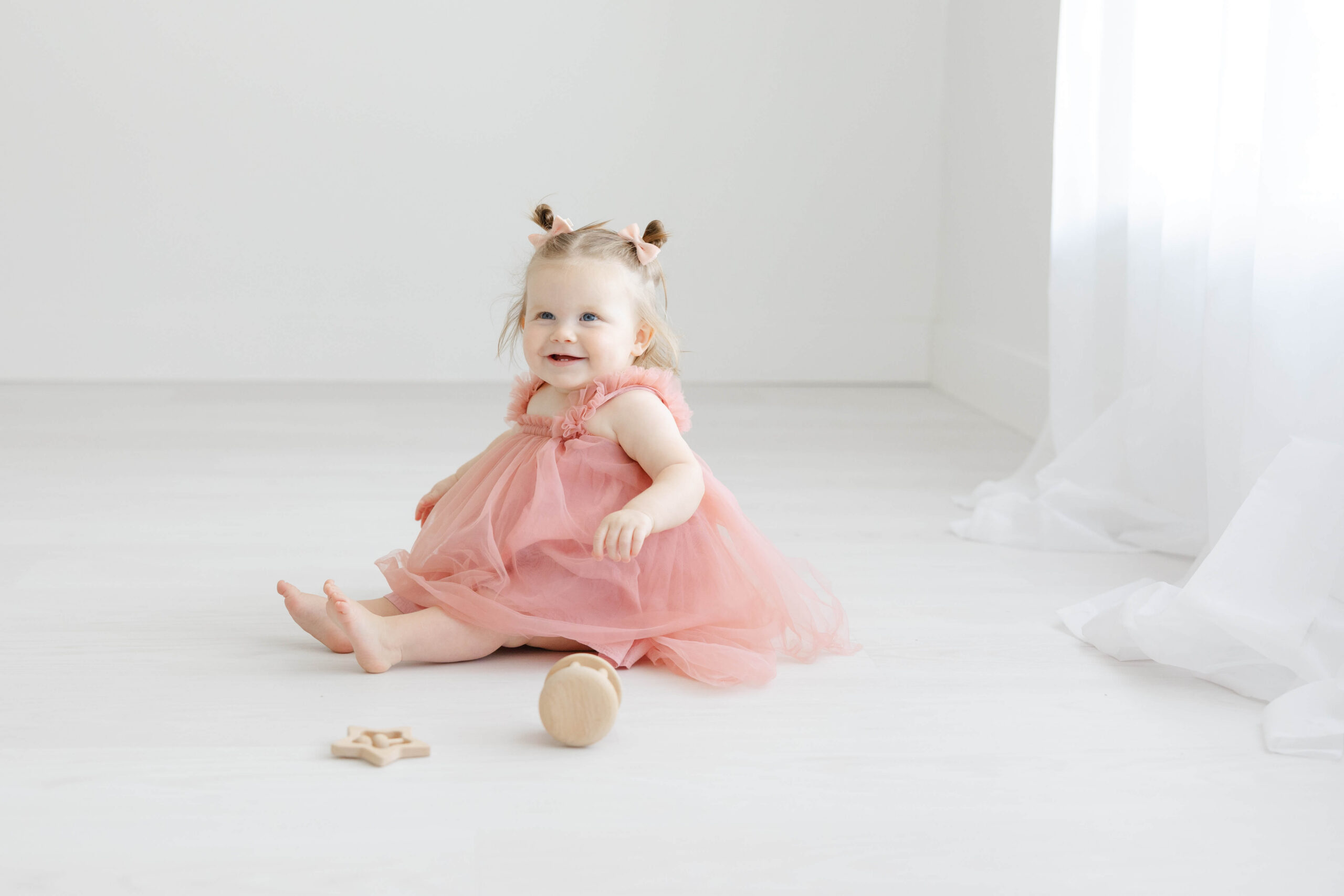 A happy young girl in a pink dress sits with some wooden toys smiling under a window after some toddler dance classes in jacksonville fl