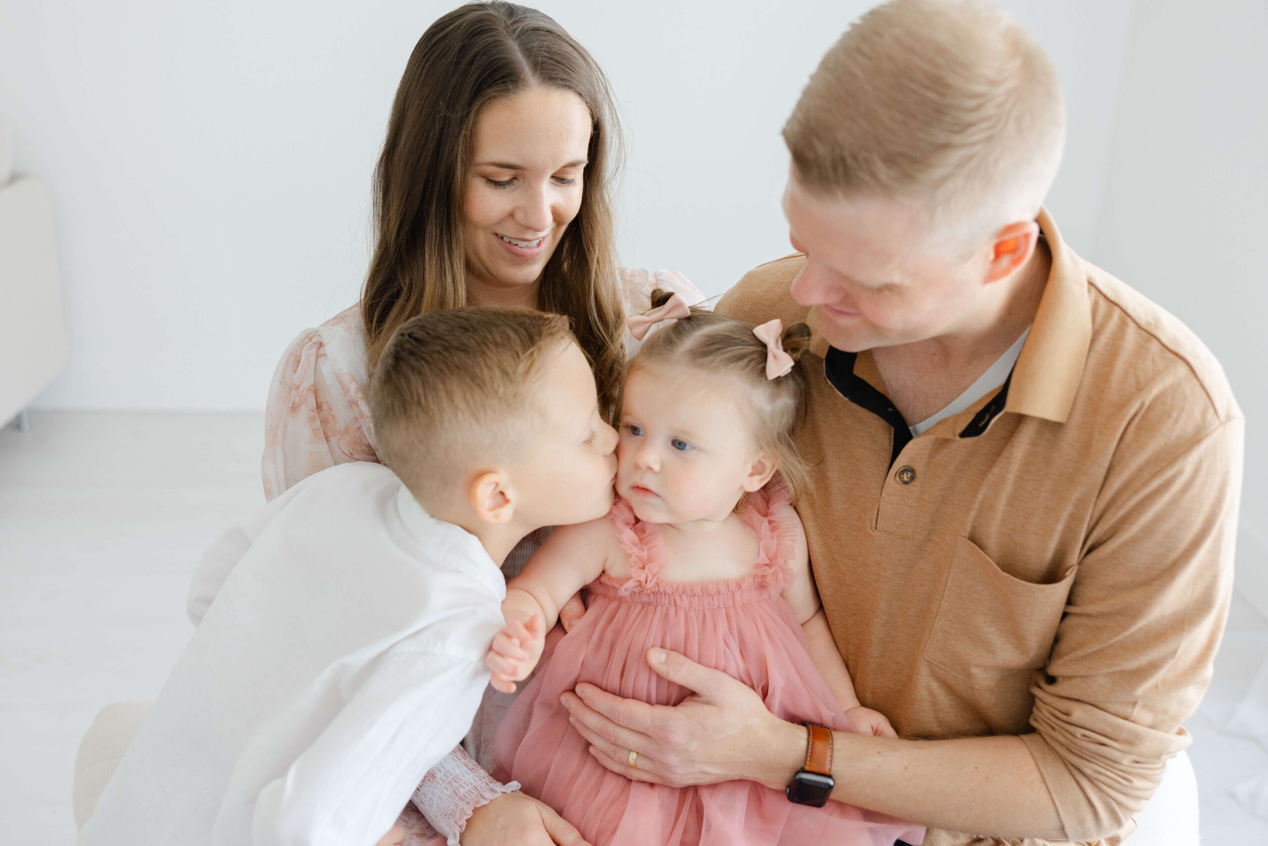a toddler boy kisses the cheek of his younger sister sitting in mom and dad's laps in a studio after some toddler dance classes in jacksonville fl
