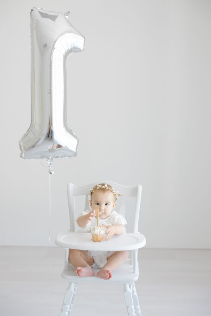 Infant photography outfit ideas for a first birthday photoshoot with a baby in a white outfit, floral crown, cupcake, and silver number one balloon.