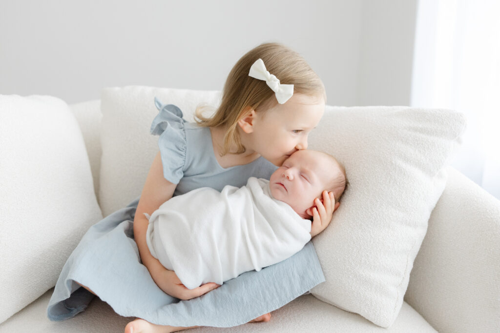 Infant photography outfit ideas featuring a baby wrapped in white while an older sister in a soft blue dress gives a gentle kiss.