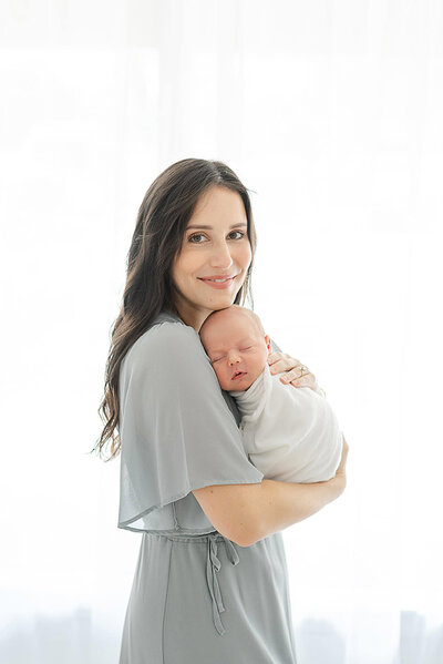 Infant photography outfit ideas with a mother in a soft gray dress holding her swaddled newborn in a bright studio.